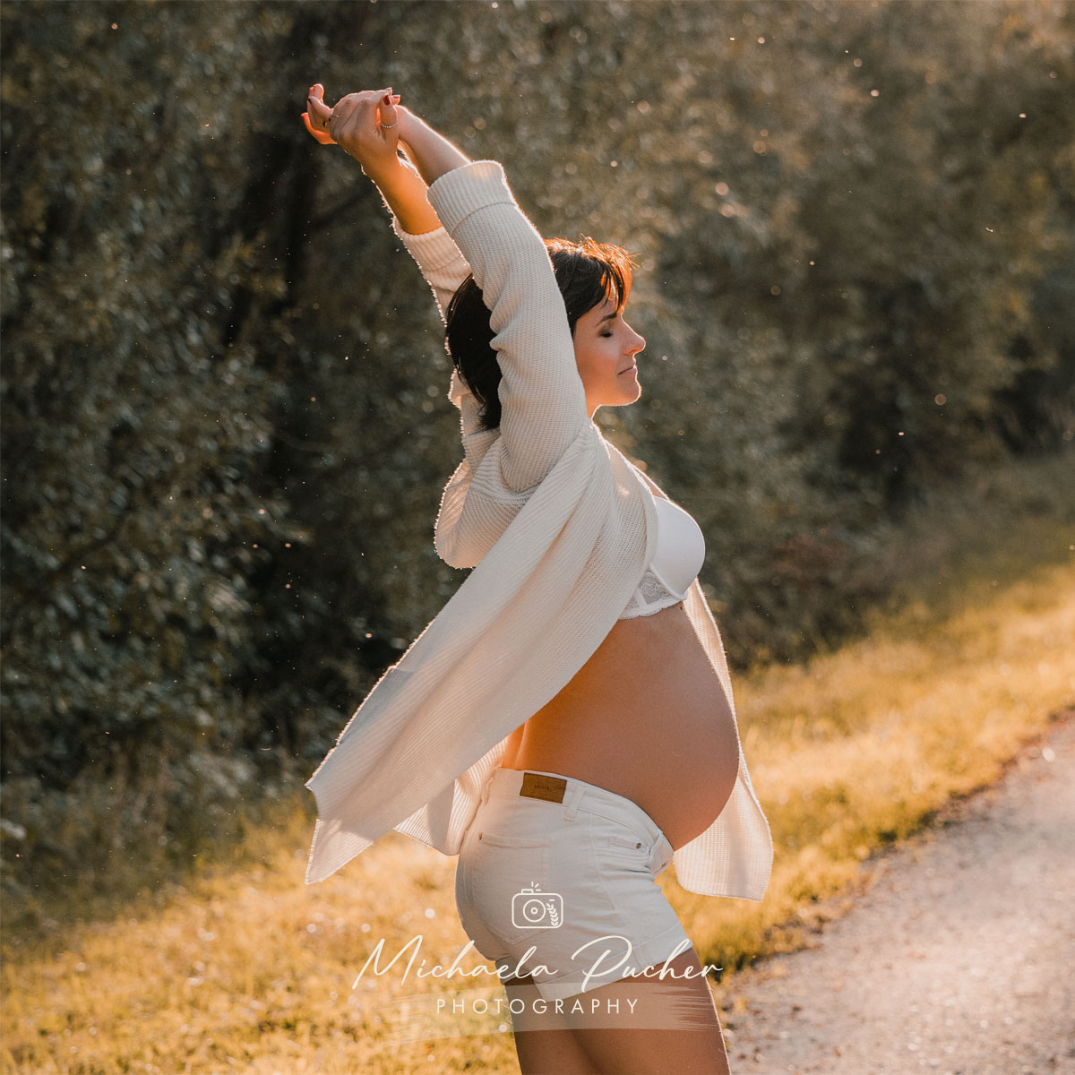 Schwangere Frau steht im warmen Abendlicht auf einem Weg, streckt entspannt die Arme nach oben und genießt den Moment; sie trägt weiße Shorts, ein weißes Top und eine offene Strickjacke.